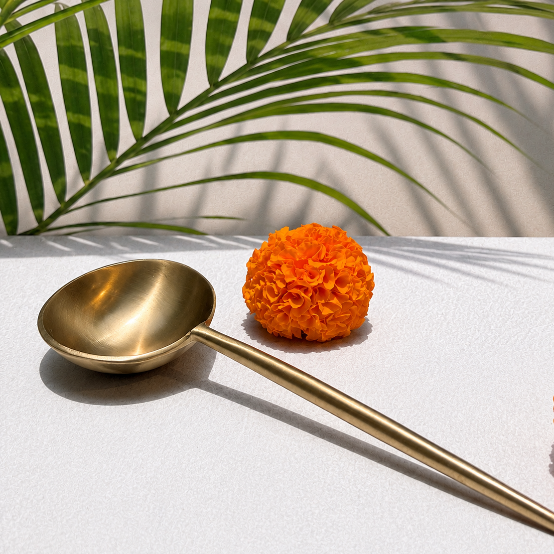 A polished brass spoon with a long handle lies on a white surface, styled with two orange marigold flowers and green palm leaves casting shadows in the background.