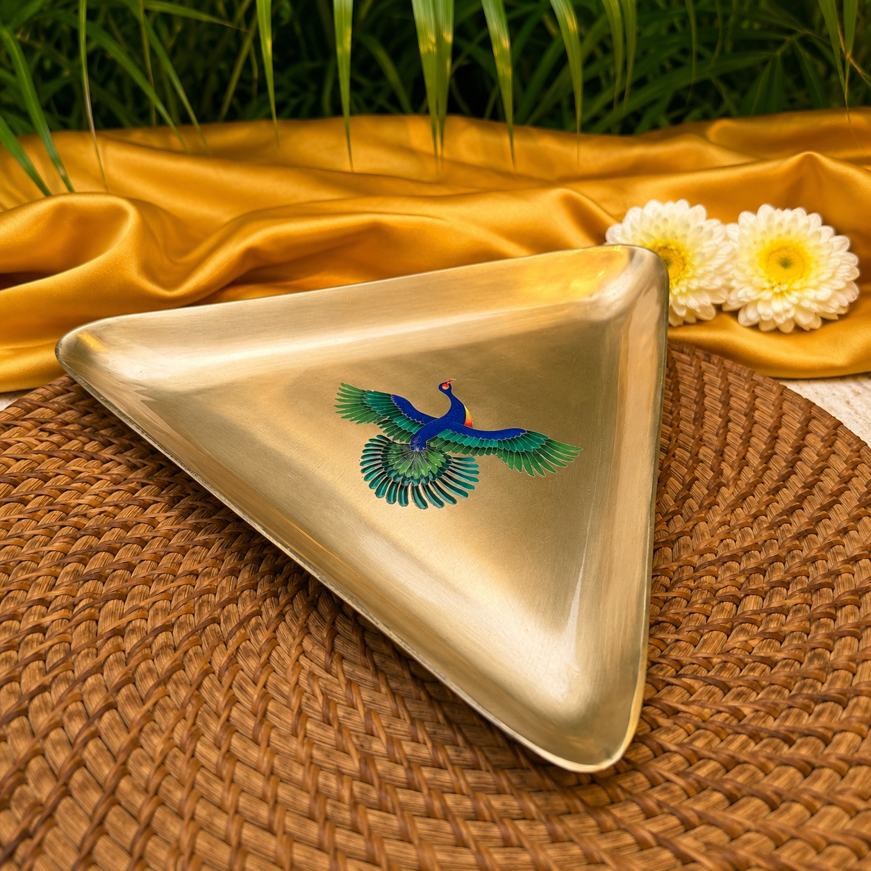 A triangular metallic tray with a polished gold finish and a painted blue peacock in the center, styled on a woven brown mat with gold fabric and white flowers in the background.