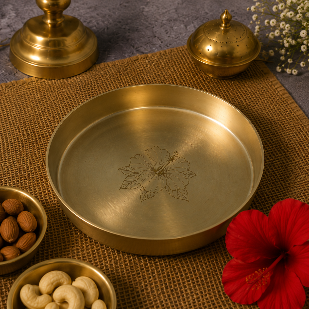A round brushed brass serving tray with an engraved floral motif in the center, styled on a brown woven mat with nuts in small bowls, a red flower, and two brass containers in the background.
