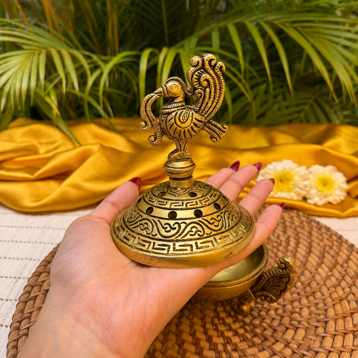 A hand holds an ornate brass incense burner with a decorative bird finial and engraved patterns, set against a backdrop of yellow fabric, palm leaves, and white flowers.
