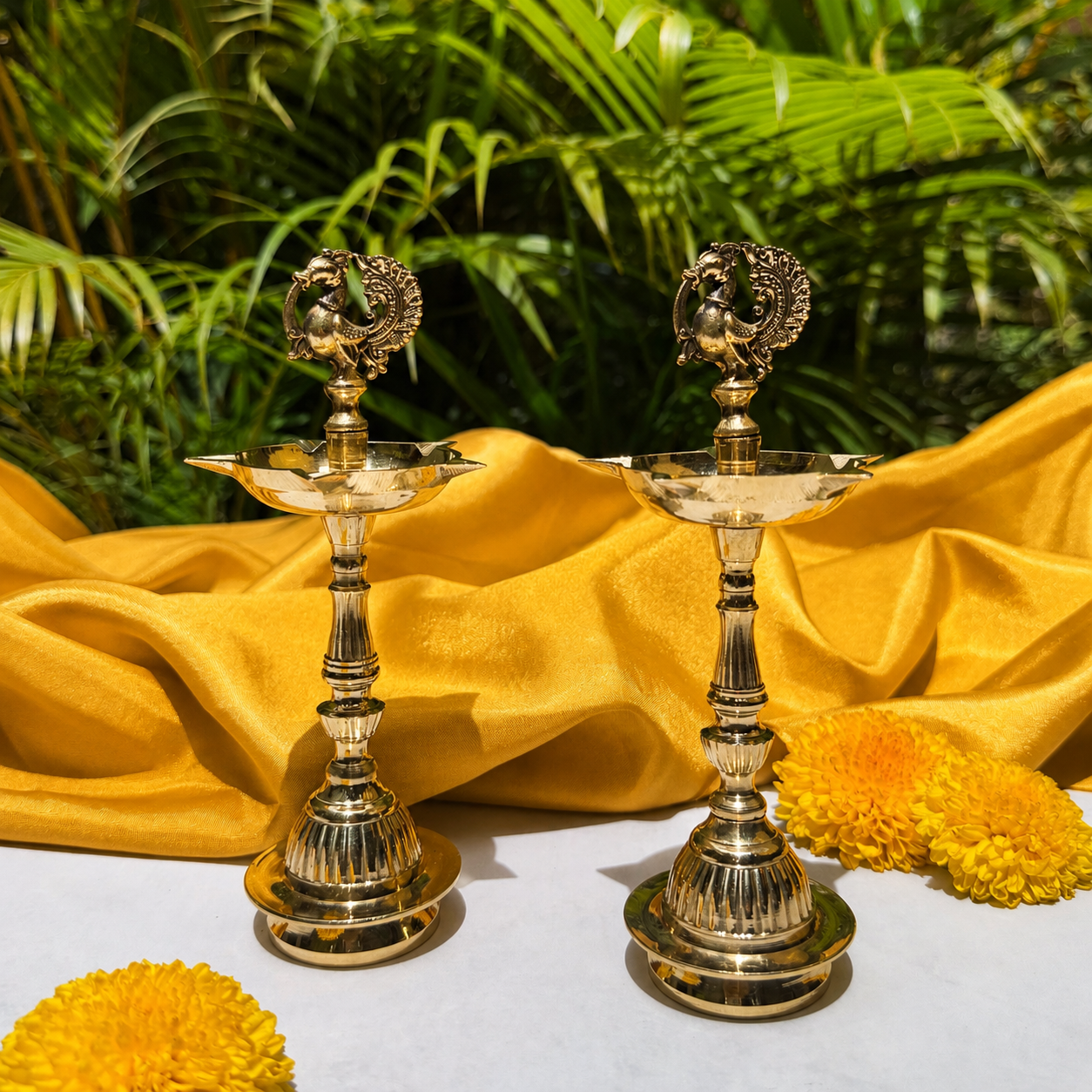 Two ornate brass oil lamps with peacock finials, styled on a white surface with yellow fabric and marigold flowers against a background of green tropical foliage.