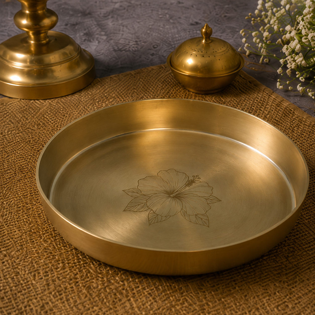 A round brass tray with a hand-engraved hibiscus flower design, displayed on a textured burlap mat with decorative brass objects in the background.