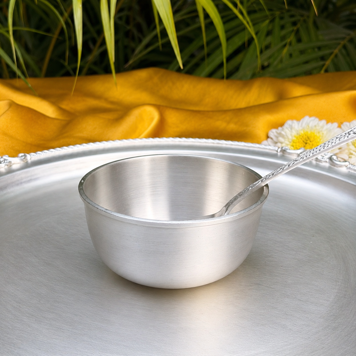 A brushed silver metal bowl with a matching spoon resting inside, set on a large silver tray. A draped yellow fabric and green leaves fill the background, with a white daisy flower on the right side.