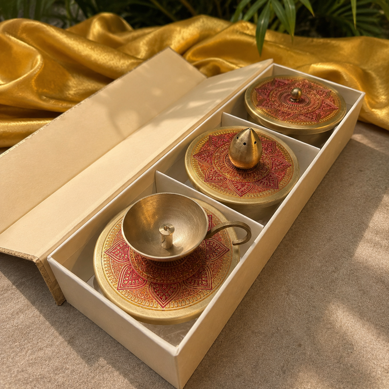 Three brass temple-style vessels with decorative red and gold patterned lids and saucers, presented in an open cream-colored gift box on a beige surface with draped gold fabric in the background.