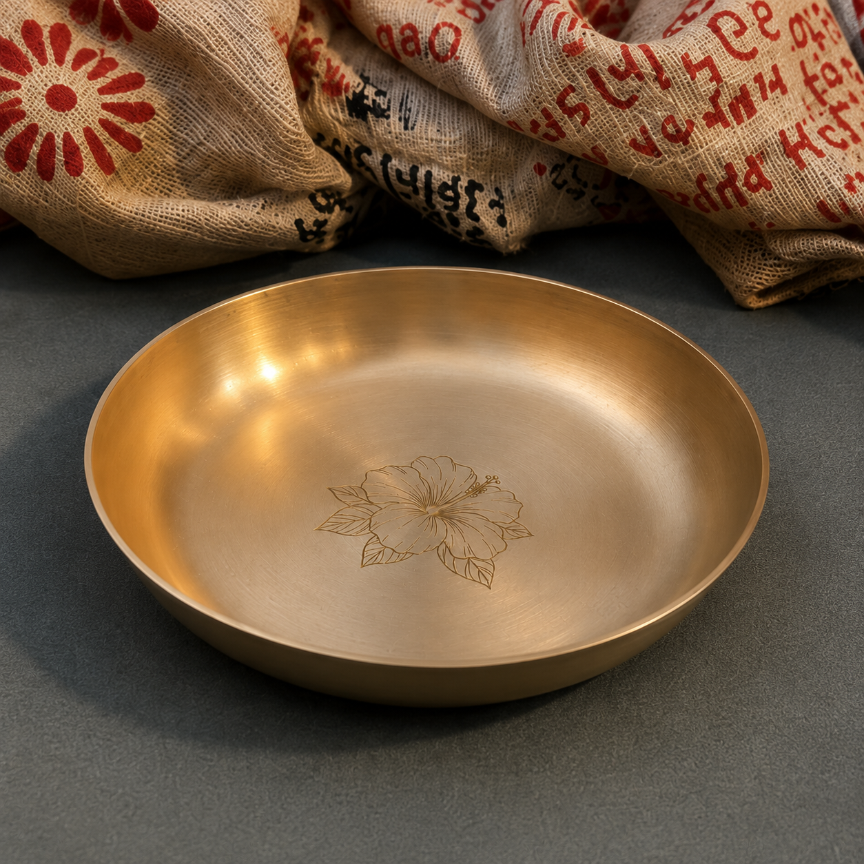 A round brass plate with a brushed metallic finish and an engraved hibiscus flower in the center, displayed on a dark gray surface with a burlap sack in the background.