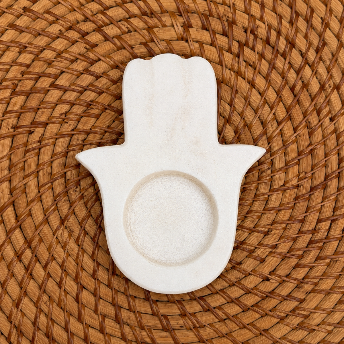 A white stone Hamsa hand tray with a circular depression in the center, placed on a round woven brown placemat.