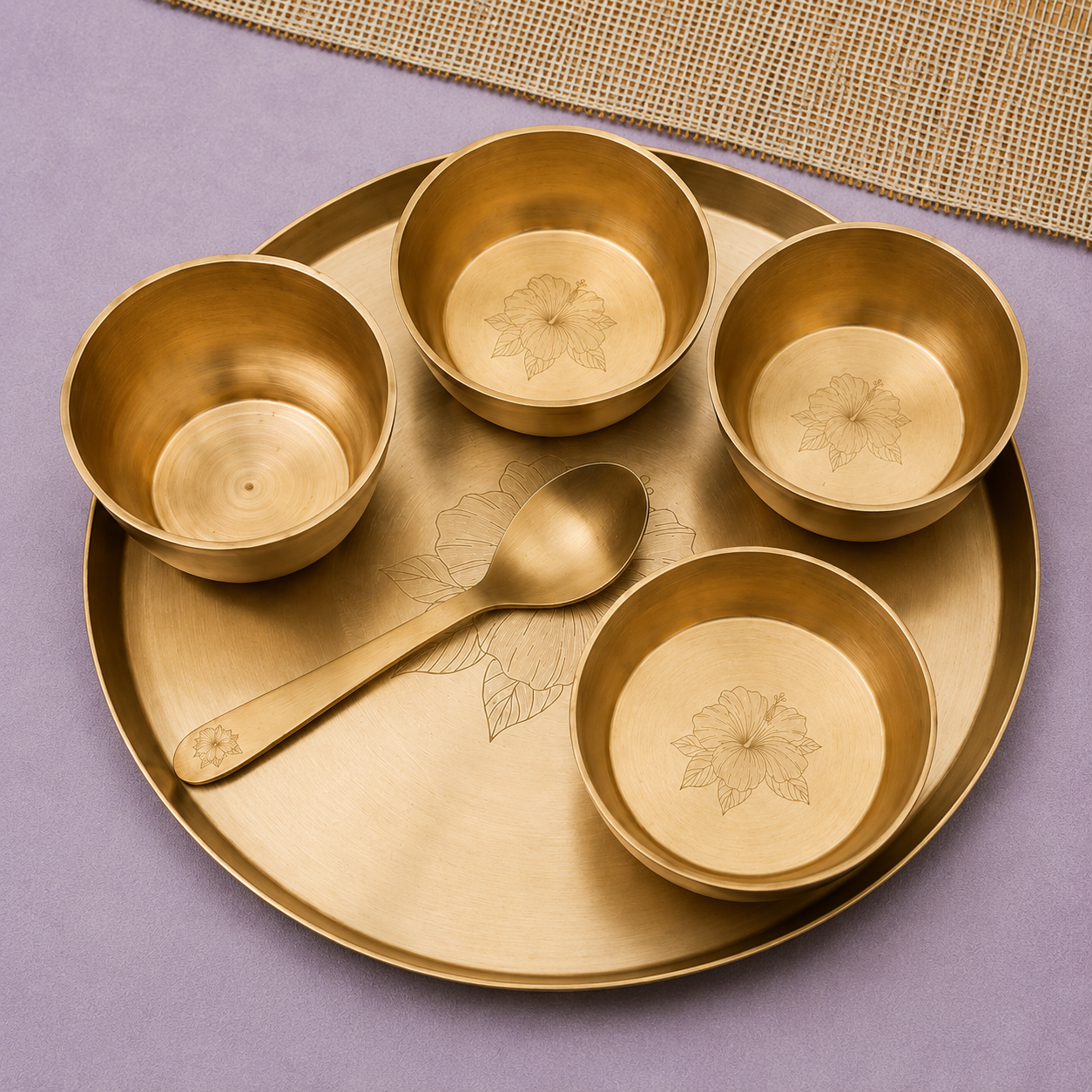 A set of four brass bowls and a matching spoon on a round brass tray, arranged on a lavender surface with a woven mat edge visible.