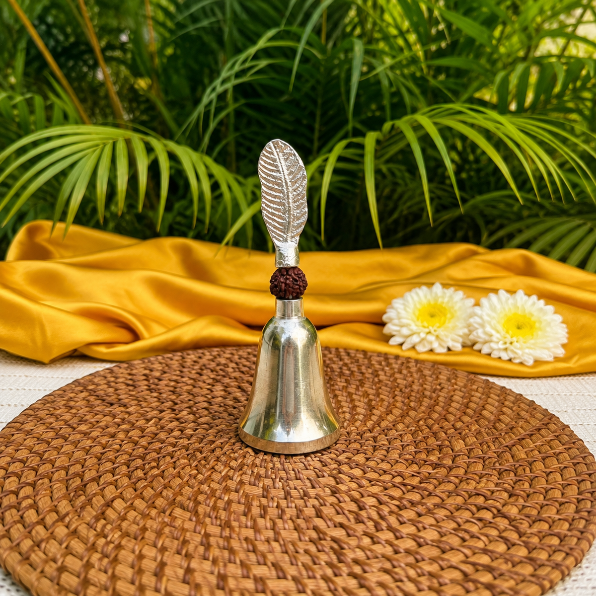 A silver-toned metal bell with a leaf-shaped handle and a dark red thread accent, displayed on a round woven brown placemat. A draped golden-yellow fabric and two white flowers sit behind it, with green palm leaves in the background.