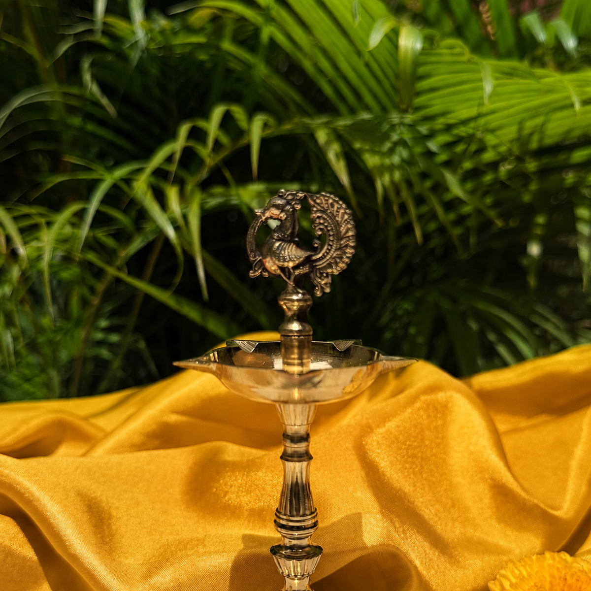 A brass oil lamp with a decorative peacock figurine on top, set on a draped golden-yellow fabric with a blurred background of green tropical leaves.