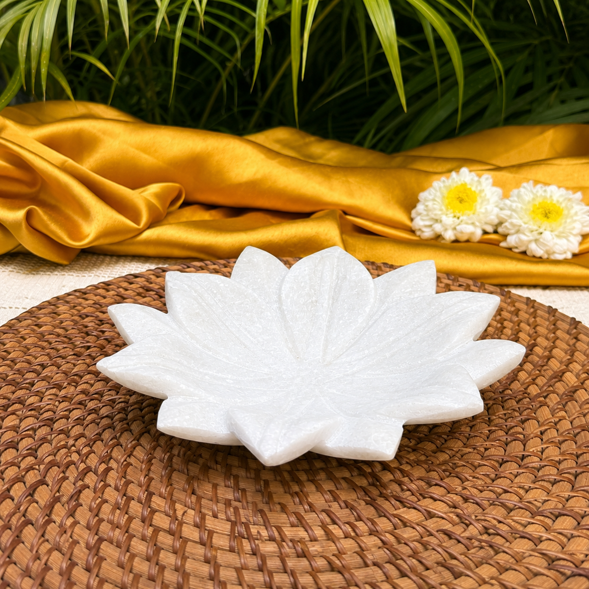 A white lotus-shaped ceramic dish or tray sits on a round woven brown placemat, with draped gold satin fabric, green leaves, and two white flowers in the background.