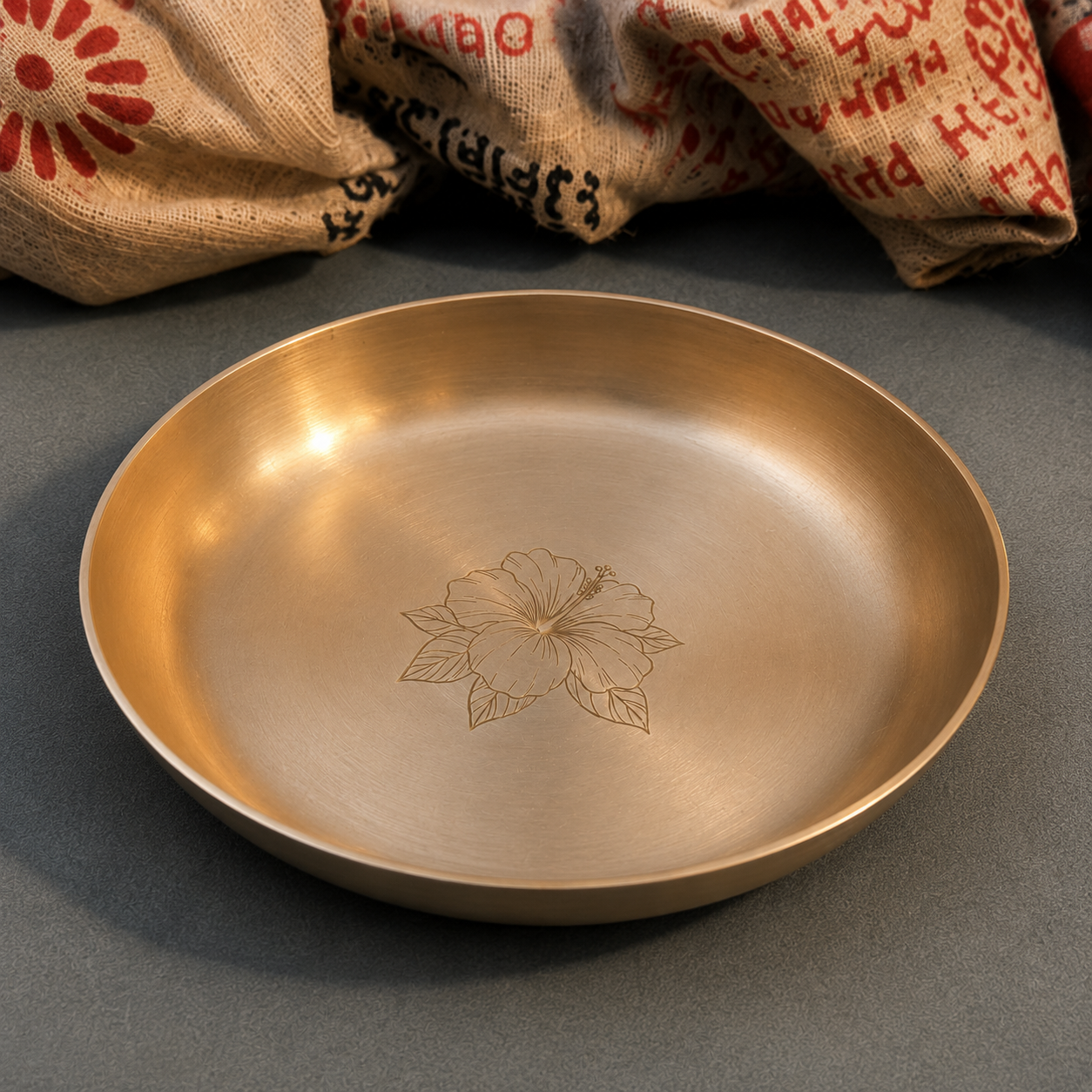 A round brushed metal tray or shallow dish with a floral engraving in the center, shown on a dark gray surface with burlap sacks in the background.