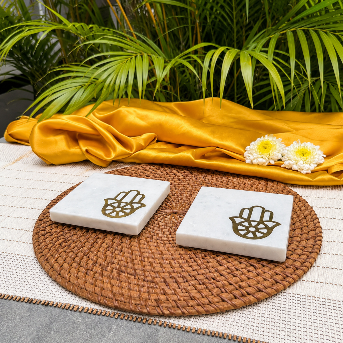 Two square white marble coasters with a gold Hamsa hand design, displayed on a round woven brown placemat with a gold draped fabric and green leaves in the background.