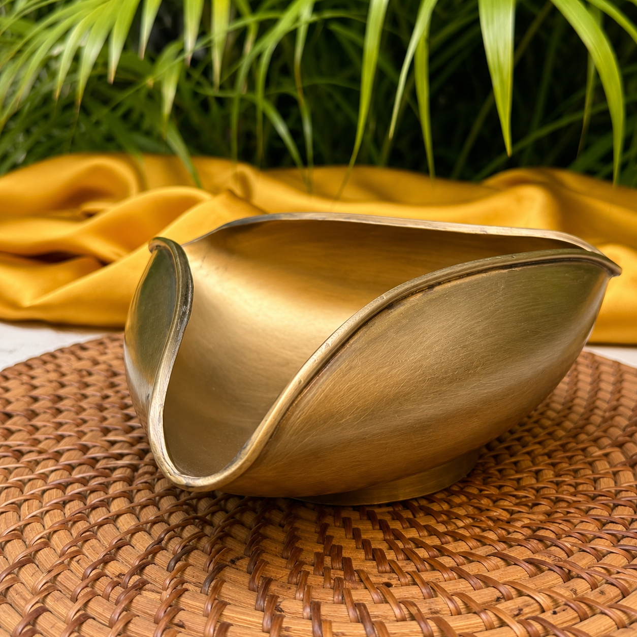 A metallic gold bowl with a unique wavy, boat-like silhouette, resting on a brown woven placemat with a draped yellow fabric and green foliage in the background.