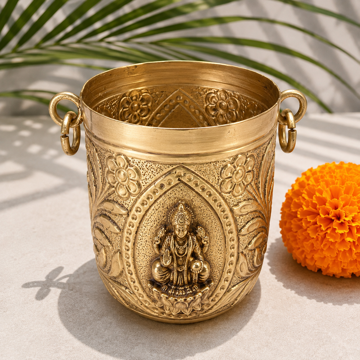 An ornate brass pot with decorative embossed floral patterns and a central deity figure, shown on a light surface with a marigold flower and palm leaves in the background.