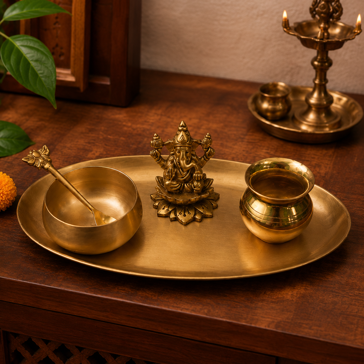 A brass Ganesha idol and matching brass tray set on a dark wooden table, alongside a brass bowl with a spoon and a small pot, with a lit brass oil lamp in the background.