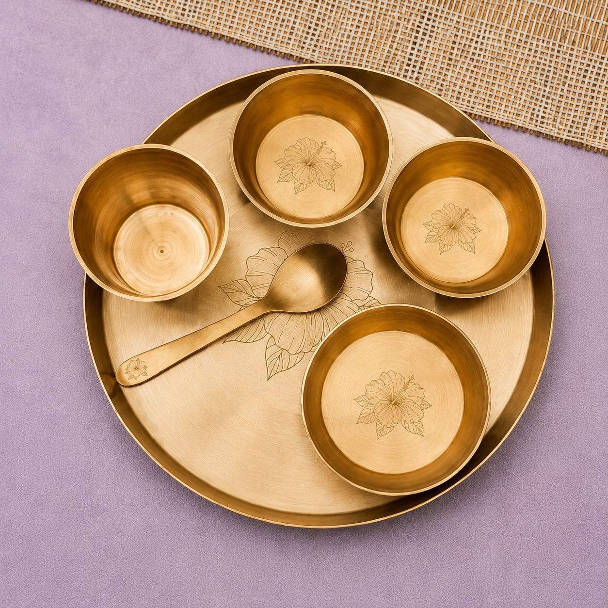 A brass plate set with a large serving platter, three small bowls, and a spoon, all decorated with floral engravings, arranged on a purple surface with a woven mat in the corner.