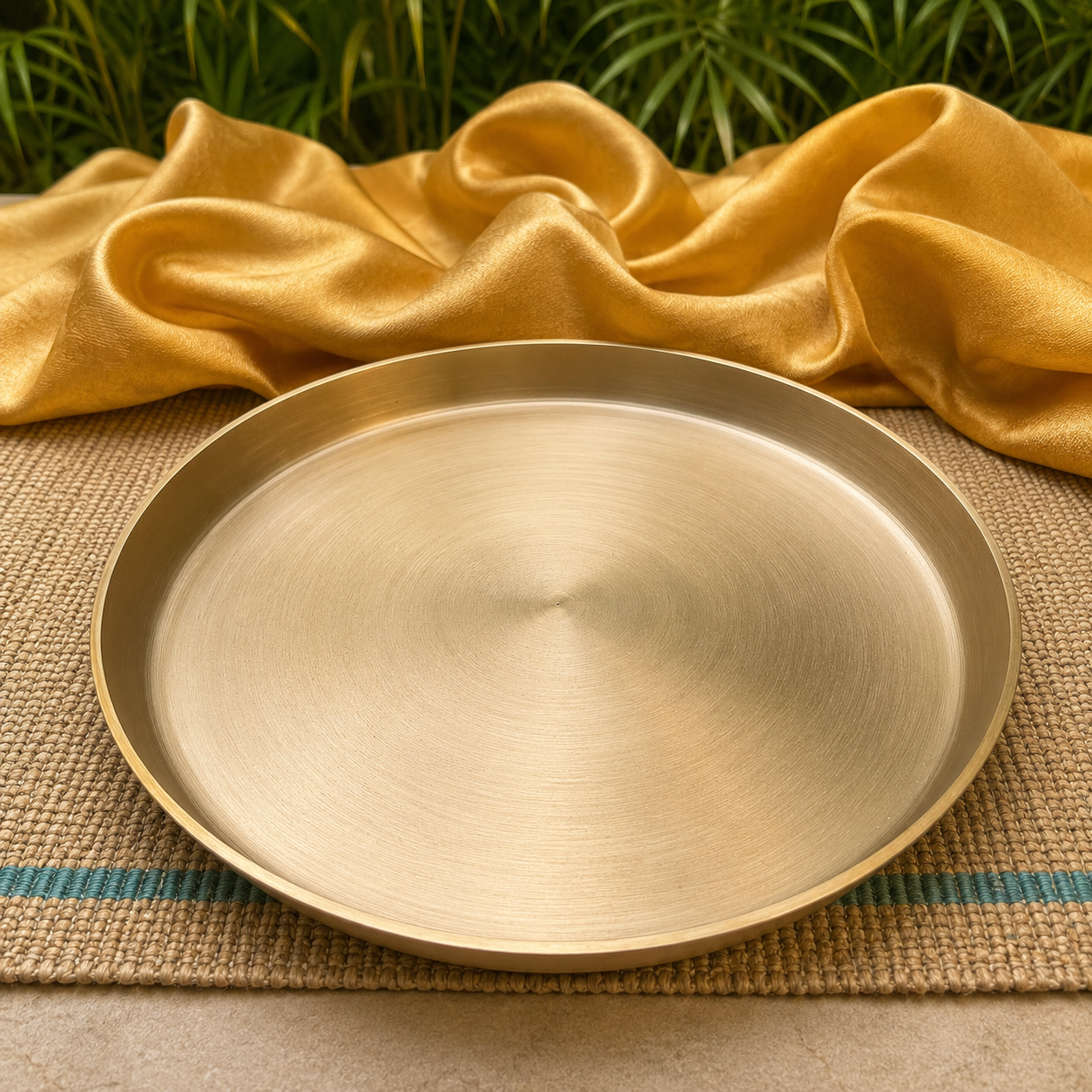 A round brushed brass serving tray or platter with a shallow rim, displayed on a woven placemat with draped gold fabric and green foliage in the background.