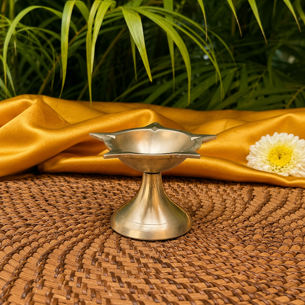 A small metallic pedestal lamp holder or diya stand with a flared base, shown on a woven brown mat with draped gold fabric and green leaves in the background.