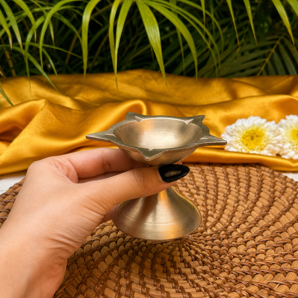 A hand with black nail polish holds a small metallic brass oil lamp (diya) with a flared, star-like rim and a pedestal base, set against a backdrop of gold fabric, white flowers, and green leaves.