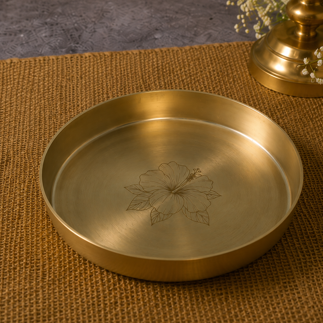 A round brass tray with a brushed metallic finish and an engraved hibiscus flower design in the center, resting on a woven brown placemat.