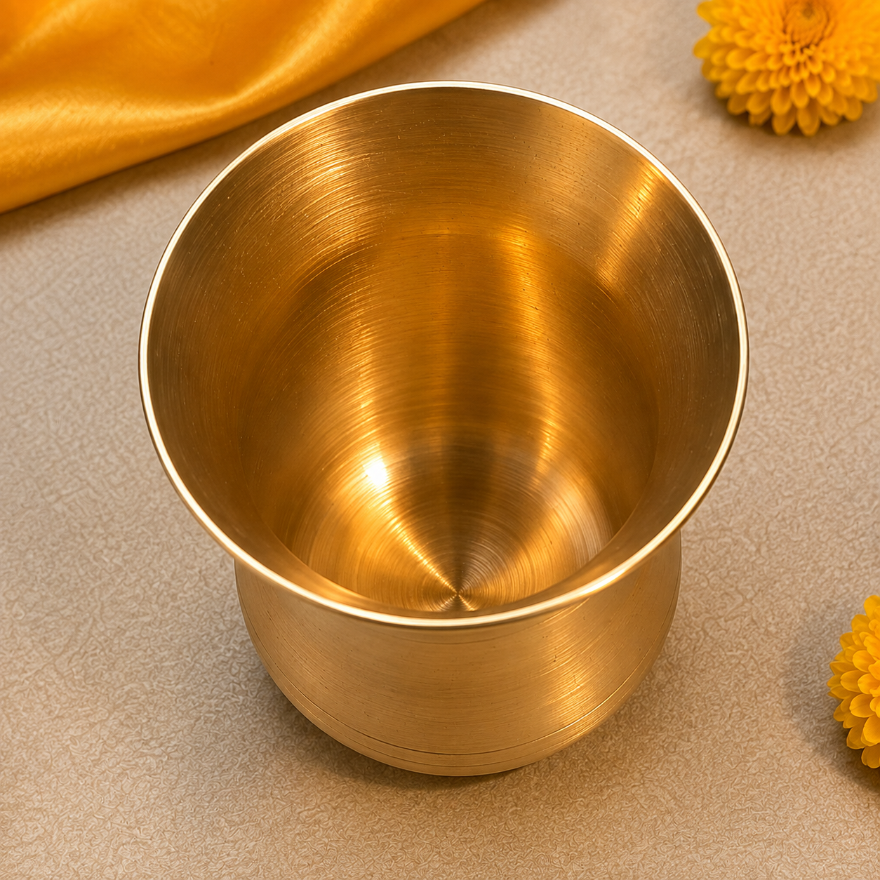A brushed brass tumbler cup with a golden finish, shown from a top-down angle on a textured beige surface with yellow flowers and a gold fabric accent.