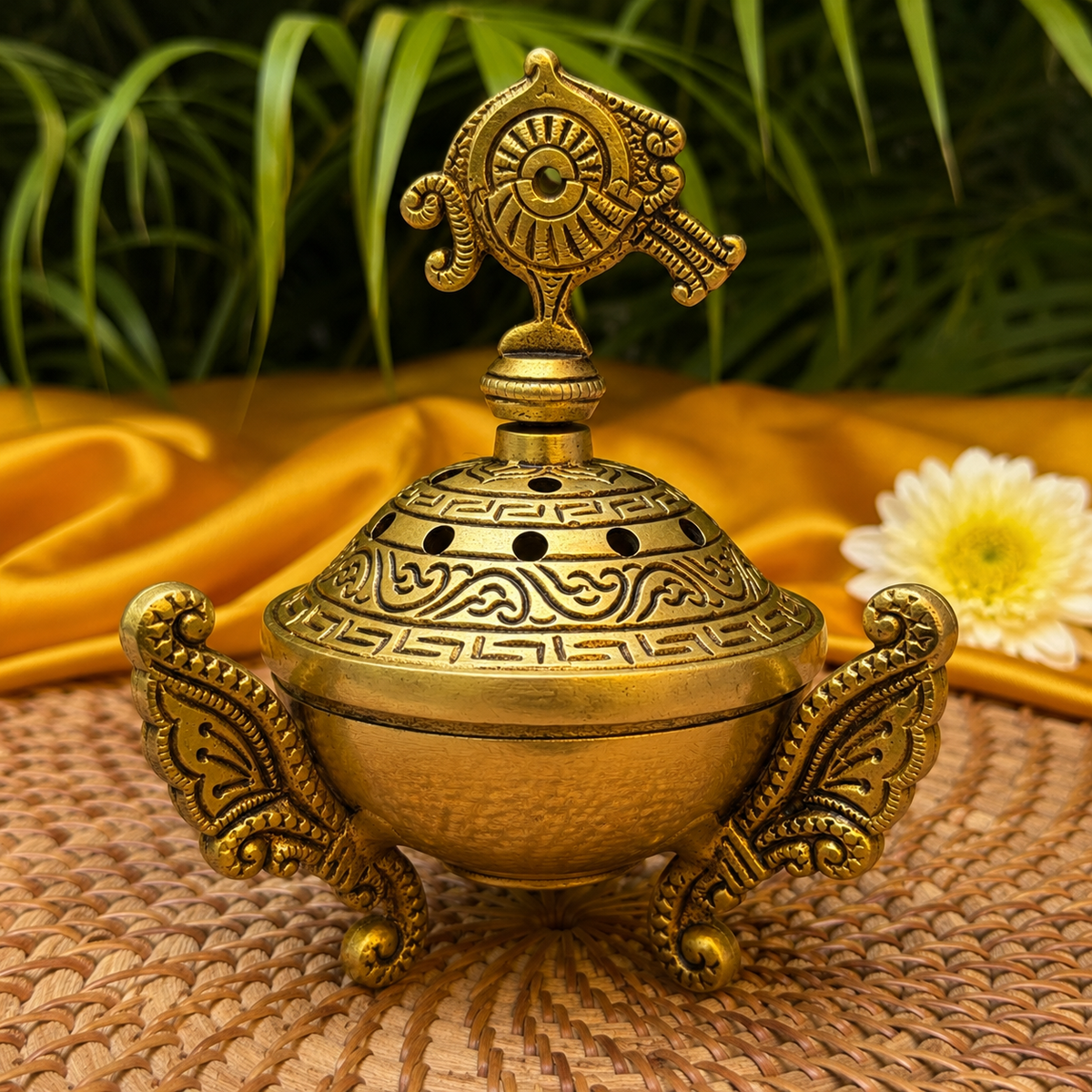 An ornate brass incense burner with intricate carvings and a decorative top handle, set on a woven mat with yellow fabric and green leaves in the background.