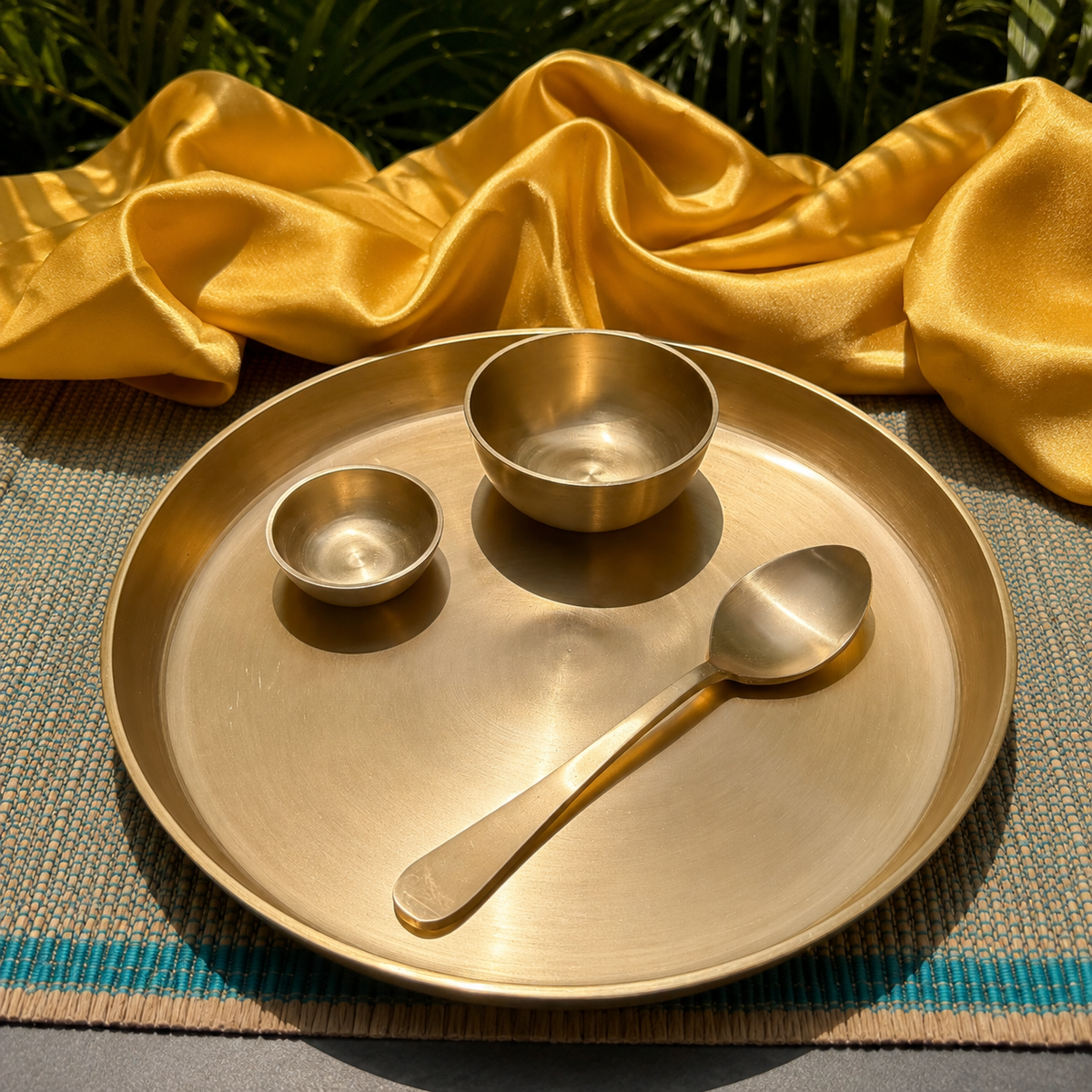 A brass serving tray with a matching small bowl and spoon, styled on a woven mat with draped gold fabric and green foliage in the background.