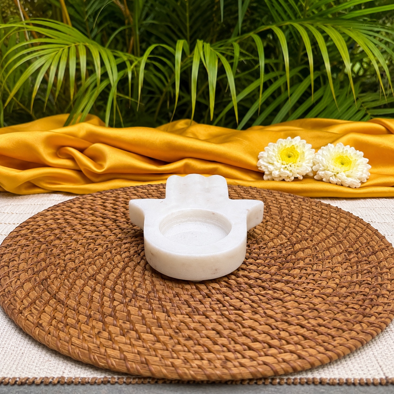 A small white stone hamsa hand holder sits on a round woven brown placemat, with a draped mustard-yellow fabric and green palm leaves in the background.