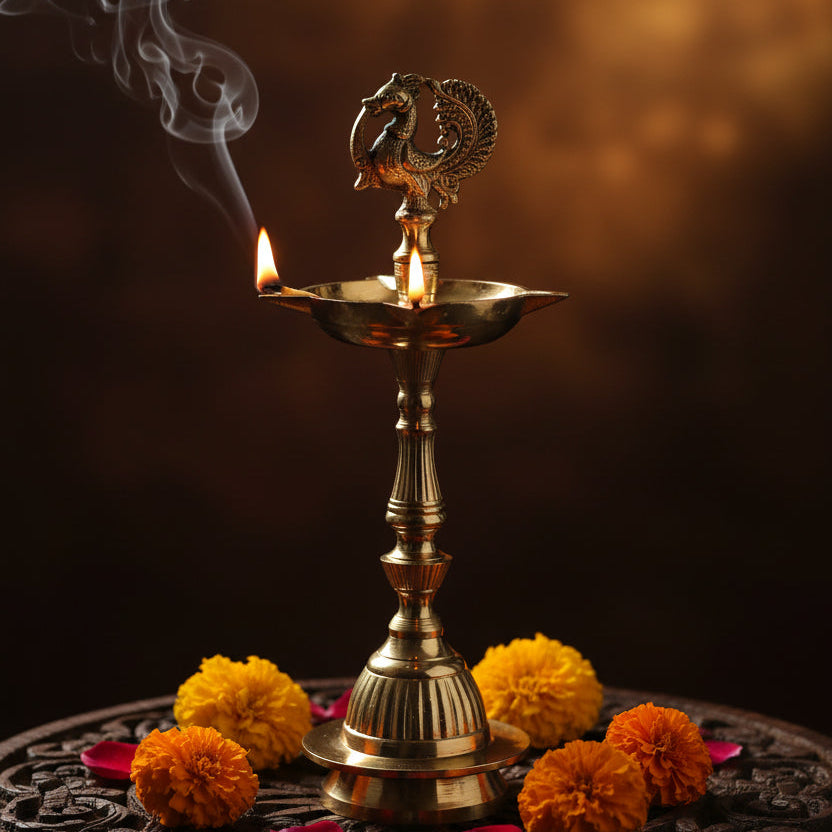 A brass oil lamp (diya) with a decorative peacock finial, set on a white surface draped with peach satin fabric and surrounded by yellow marigold flowers, with a blurred green foliage background.