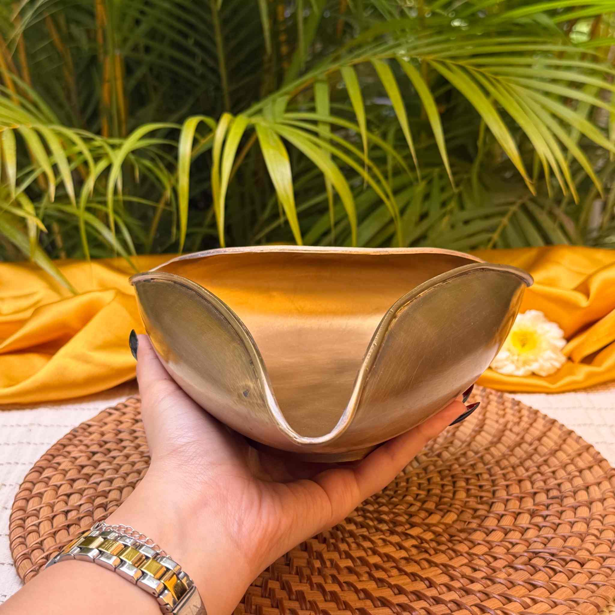 A hand holds a metallic gold-toned decorative bowl with a wavy, scalloped rim, set against a background of green palm leaves, yellow fabric, and a woven placemat.