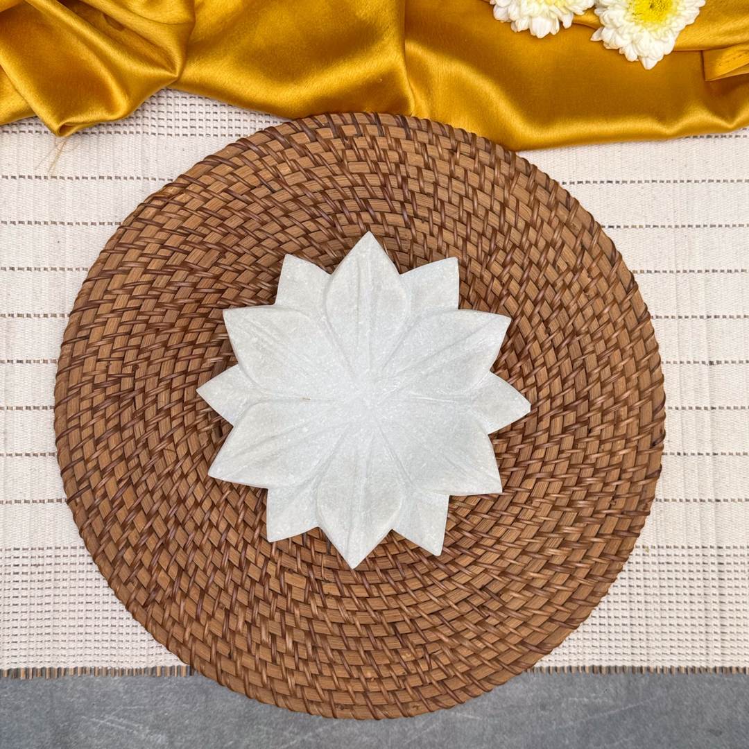 A round woven brown placemat or tray sits on a white textured table runner, with a white ceramic star-shaped plate in the center and draped gold satin fabric with white flowers at the top.