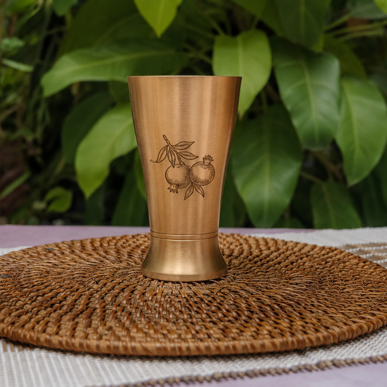 A brushed brass or bronze drinking cup with an engraved pomegranate design, displayed on a round woven placemat with a background of green foliage.