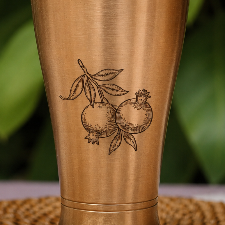 A close-up of a brushed metallic tumbler or cup with an engraved pomegranate illustration on the front, set against a blurred green background.