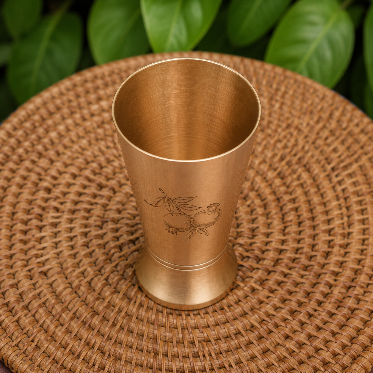 A brushed brass cocktail muddler with an engraved dragon design, resting on a round woven straw placemat with green leaves blurred in the background.