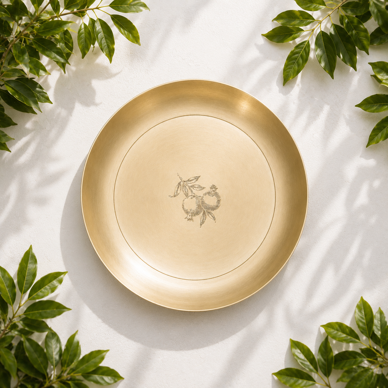 A round gold-toned serving platter with a delicate line design and a small fruit illustration in the center, styled on a white surface with green leaves in the corners.
