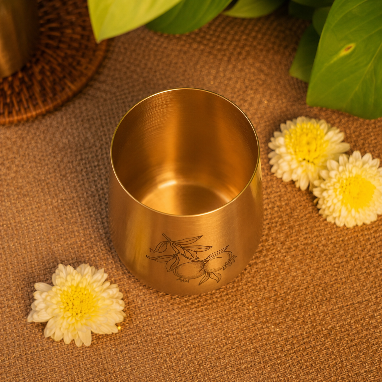 A small metallic gold cup with a hand-drawn bee engraving, placed on a brown burlap surface with white flowers and green leaves in the background.