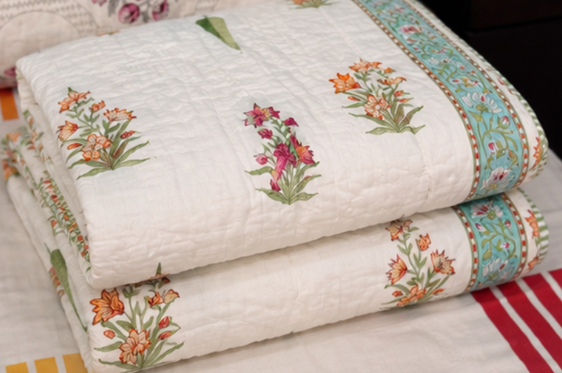 A stack of folded white quilted blankets or quilts with embroidered floral motifs in orange, pink, and green, plus a decorative turquoise border, shown on a bed.