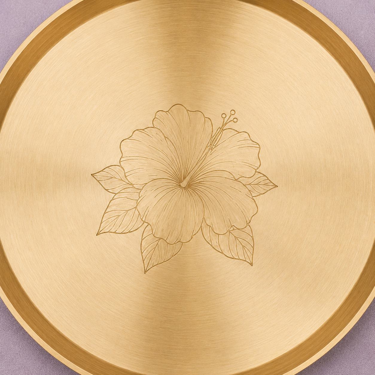 A round brushed brass tray with a laser-engraved hibiscus flower in the center, shown against a soft lavender background.