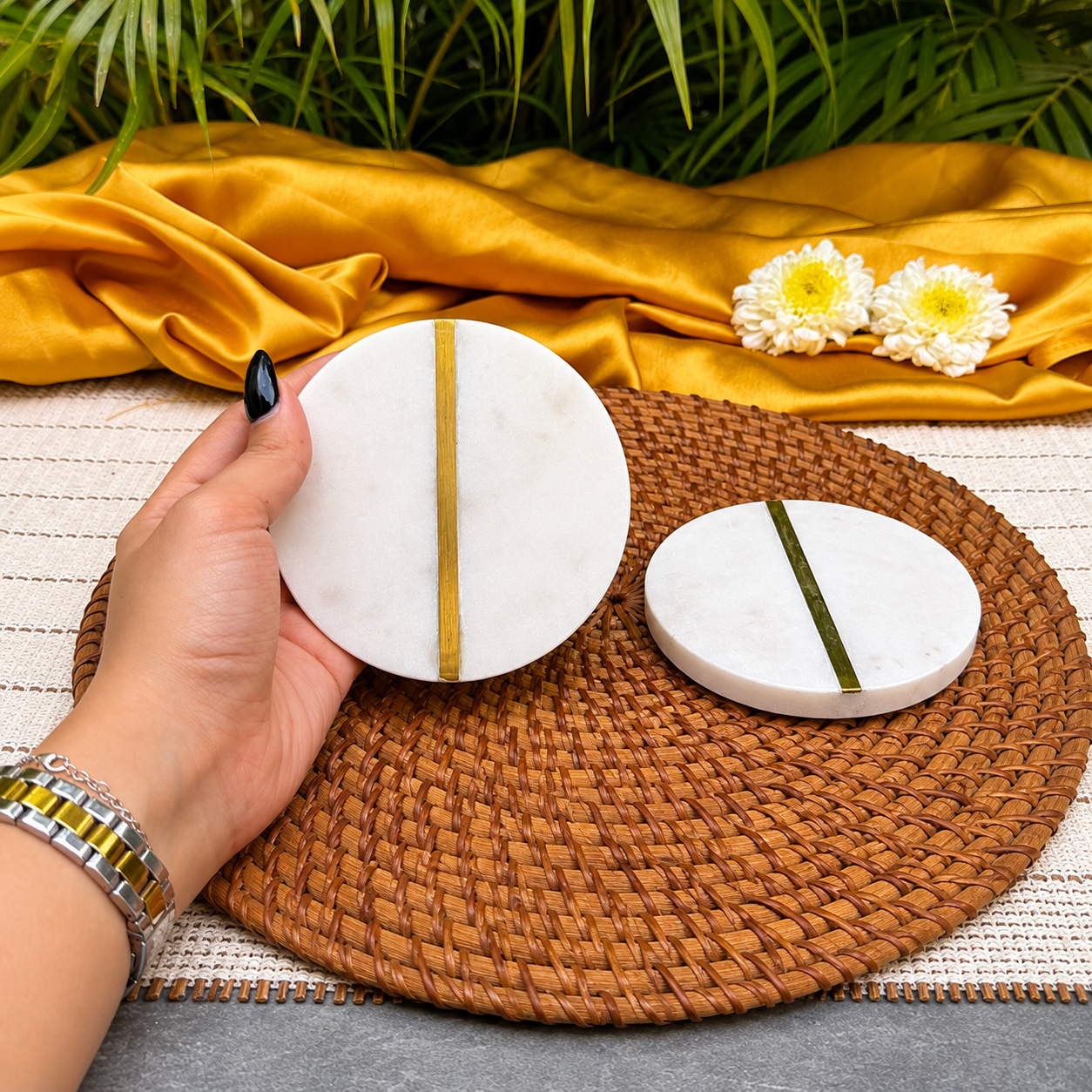 Two round white marble coasters with a vertical gold stripe, shown on a woven brown placemat with a hand holding one coaster. A gold satin cloth, green leaves, and white flowers are in the background.
