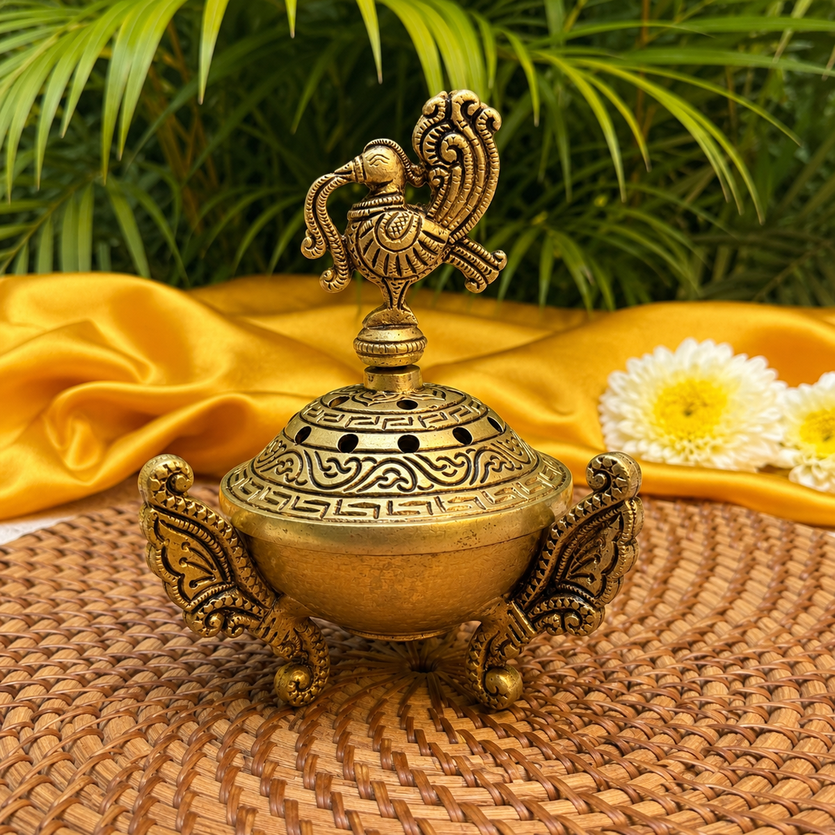 An ornate brass incense burner with a decorative bird finial on top, set on a woven mat with yellow fabric and green foliage in the background.