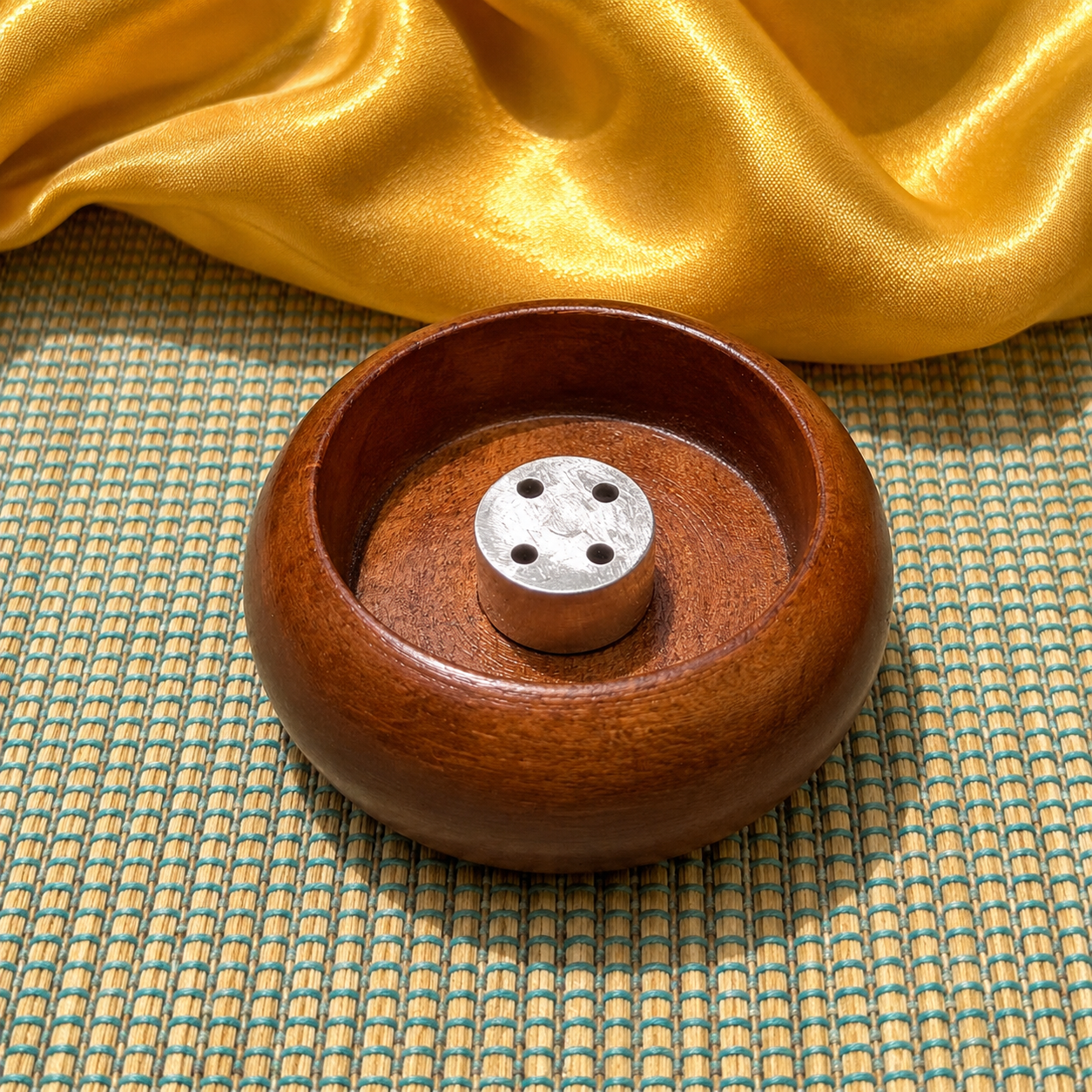 A round wooden incense holder with a small metal incense stick burner in the center, placed on a woven mat with draped gold fabric in the background.