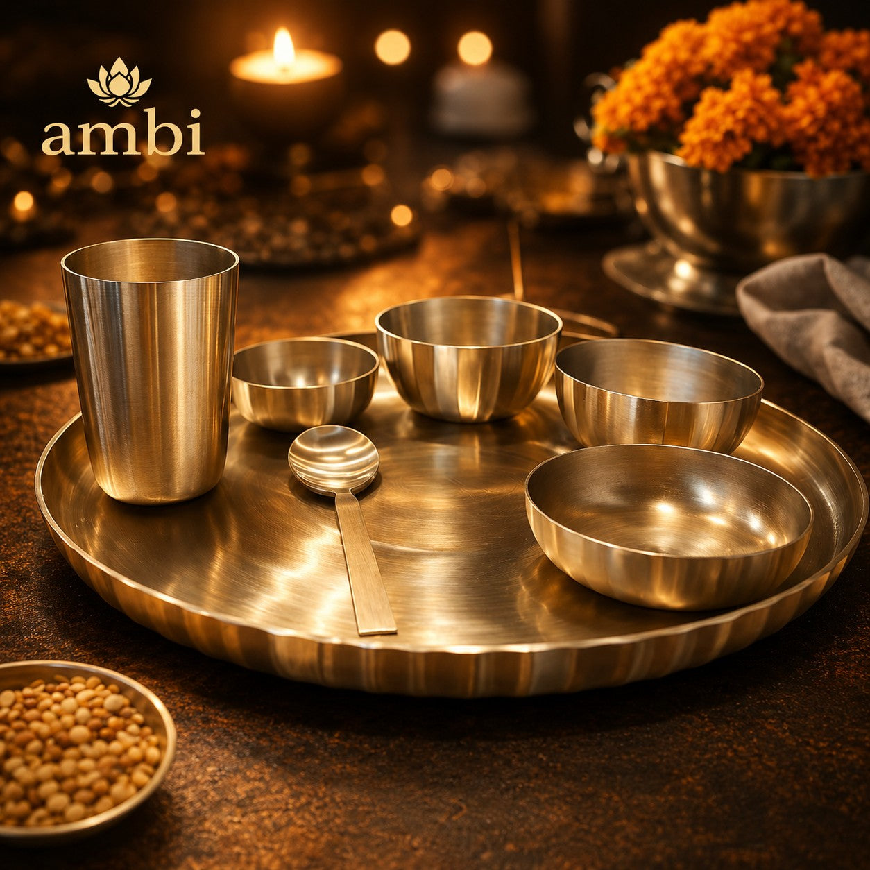 A set of metallic Ambi stainless steel tea and coffee cups and bowls arranged on a large round brass tray, with a spoon, set in a warm, dimly lit environment with candles and flowers.