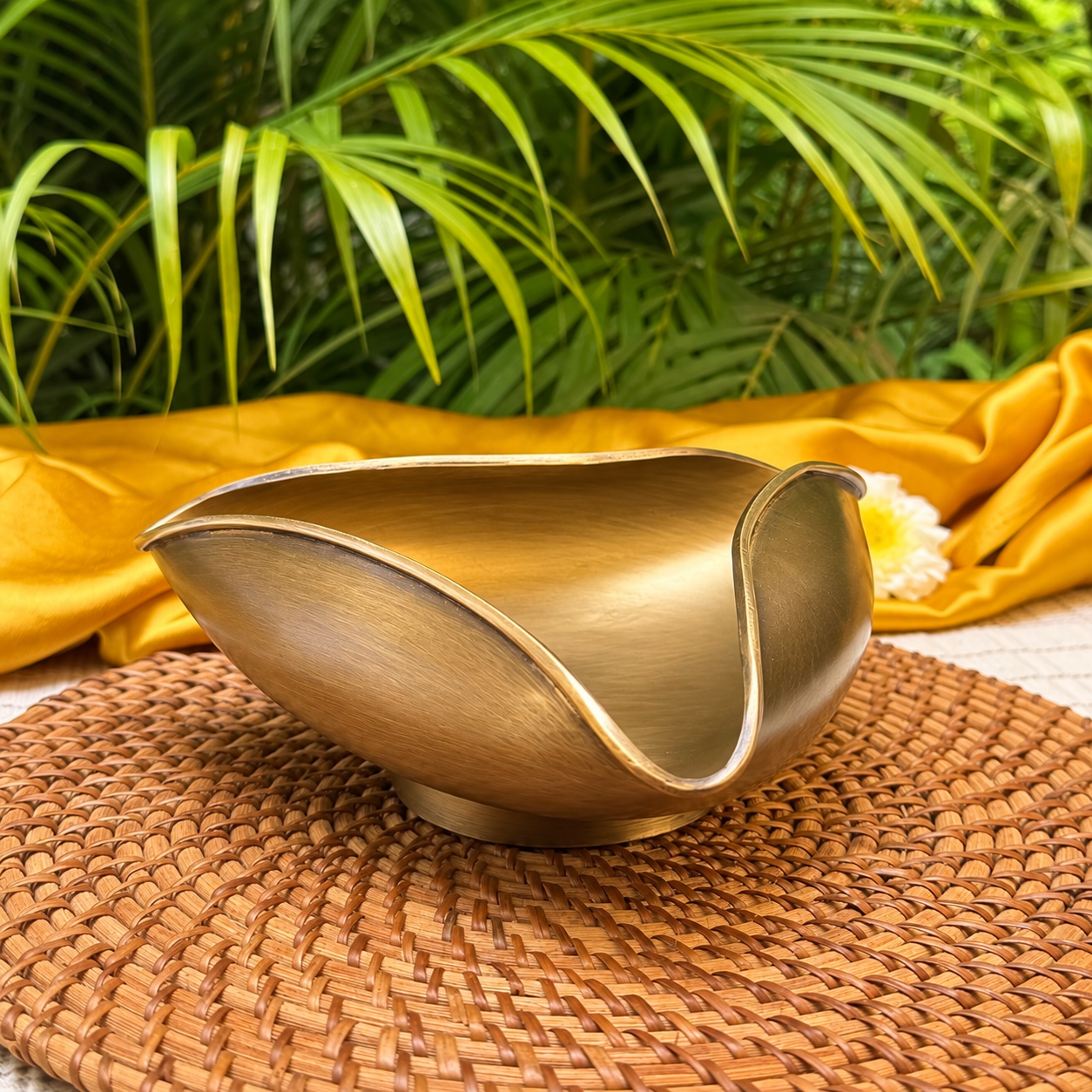 A brushed brass or bronze serving bowl with a wide, flared rim sits on a woven brown placemat, with draped yellow fabric and green palm leaves in the background.