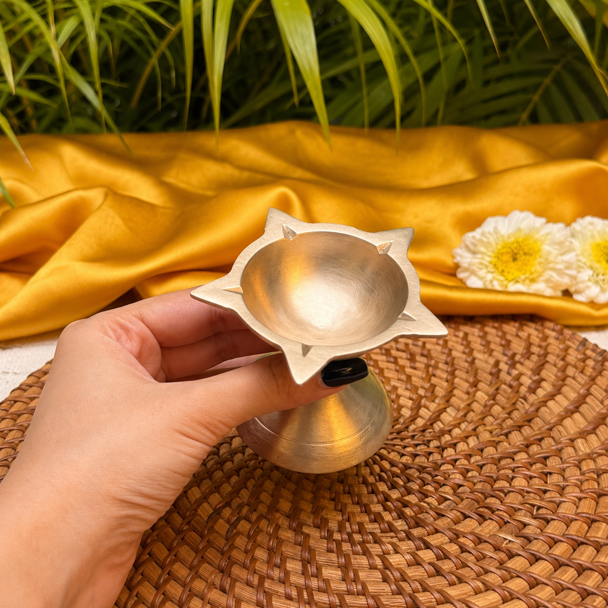 A hand holds a small metallic brass oil lamp (diya) with a star-shaped rim and a conical base, set against a background of draped golden silk fabric, white flowers, and green leaves.