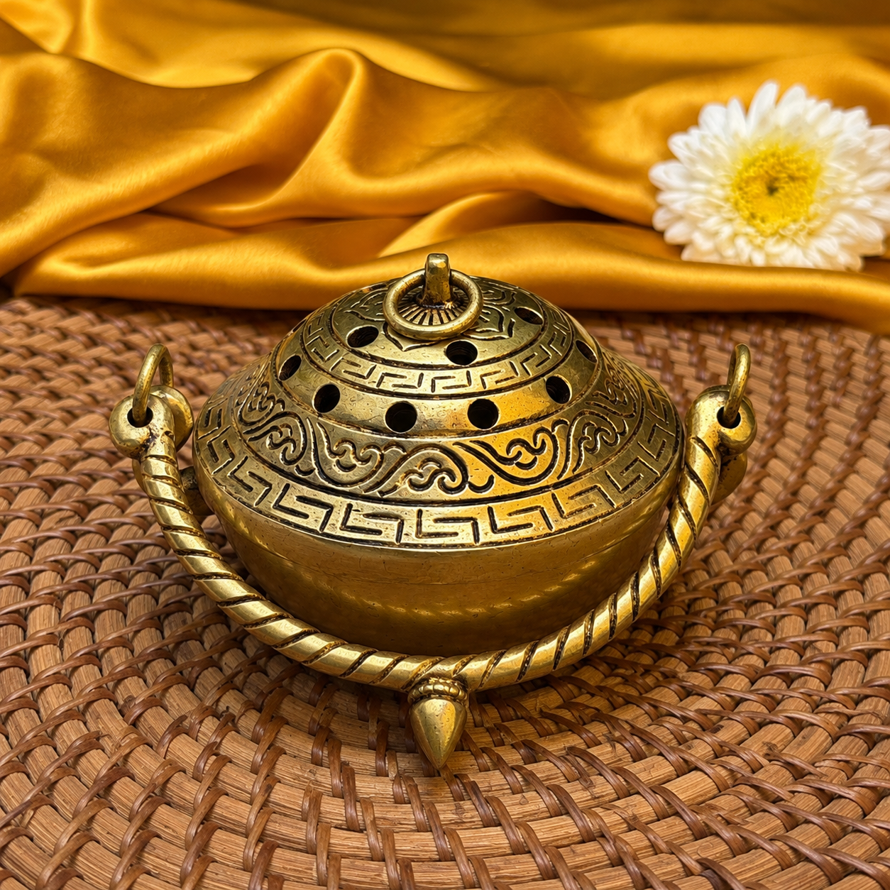 An ornate brass incense burner with carved Greek key patterns and a twisted handle, resting on a woven brown mat with gold satin fabric and a white daisy in the background.