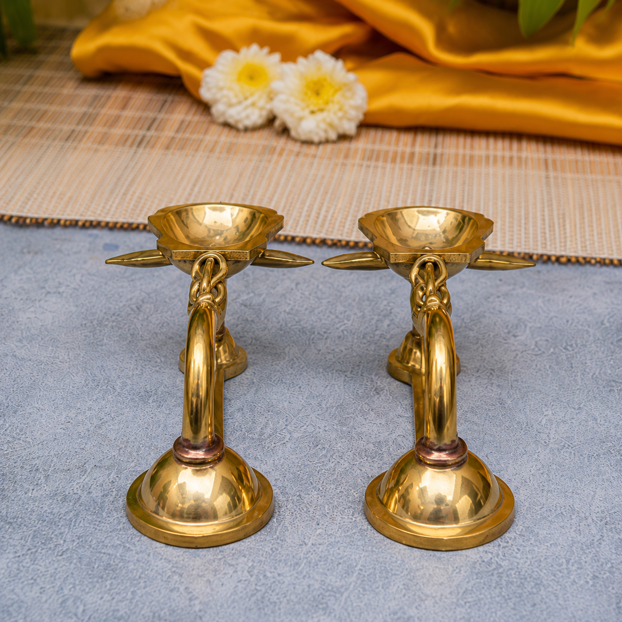 A pair of polished brass oil lamps with decorative handles and saucers, displayed on a textured blue-gray surface with a yellow satin cloth and white flowers in the background.