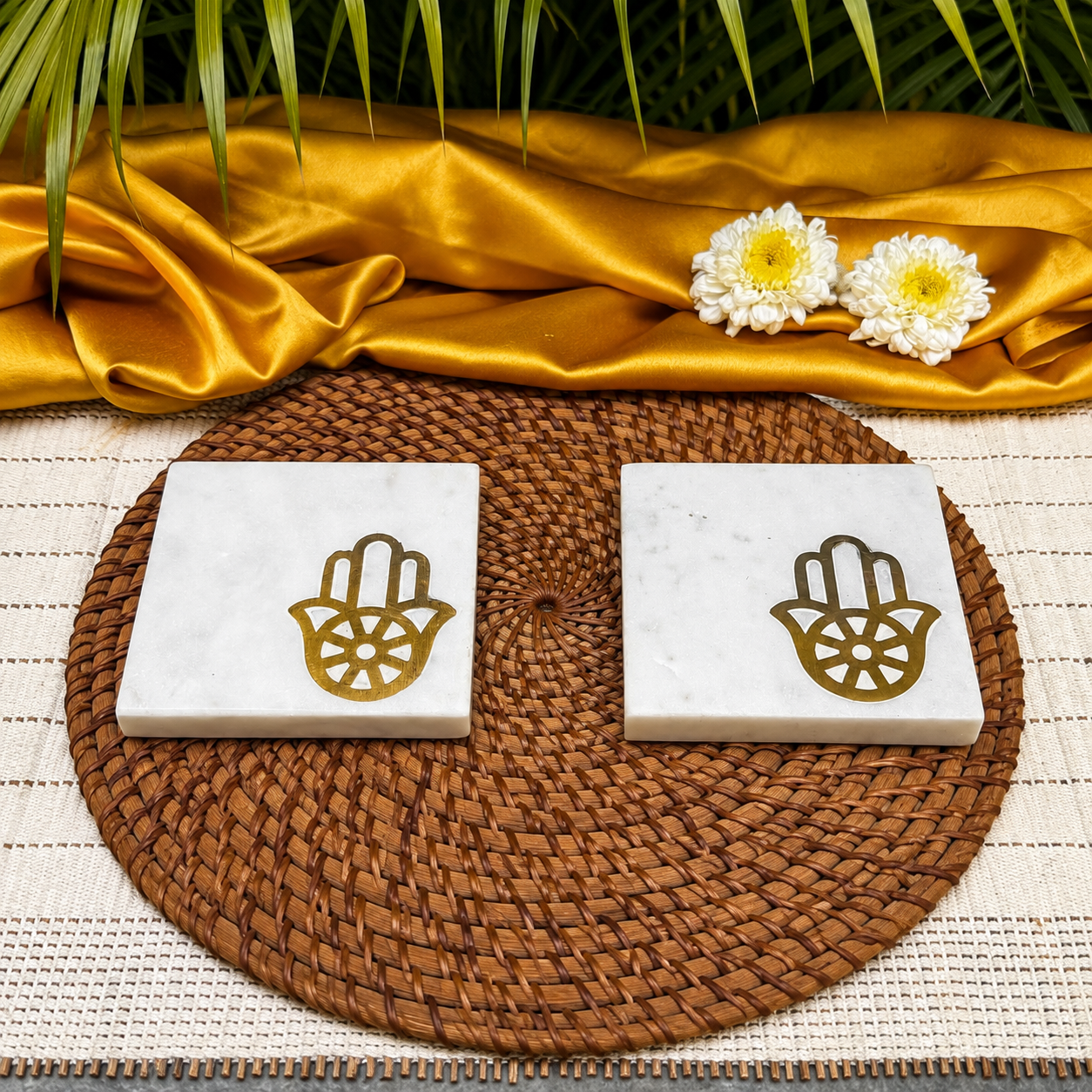 Two square white marble coasters with gold Hamsa hand designs, arranged on a round brown woven placemat with gold fabric and white flowers in the background.
