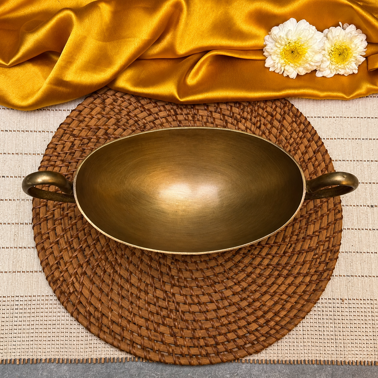 An oval bronze serving bowl with two side handles, displayed on a round woven placemat with draped gold satin fabric and two white flowers in the background.