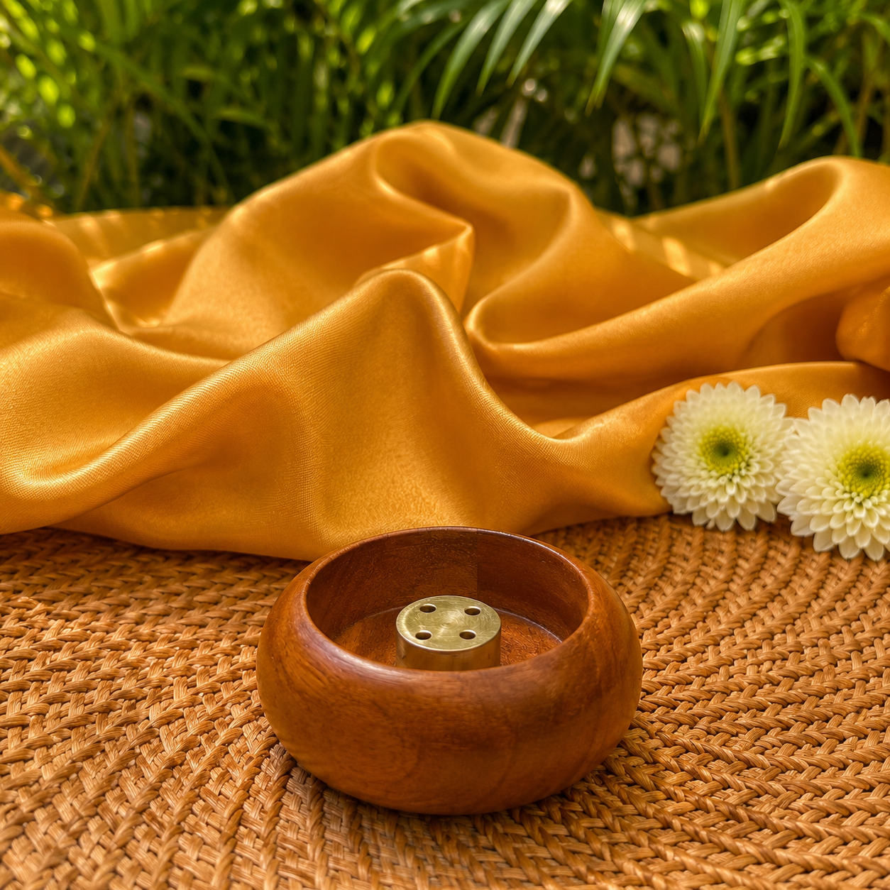 A small round wooden incense holder with a brass incense burner inside, placed on a woven straw mat with draped golden-yellow fabric and white flowers in the background.