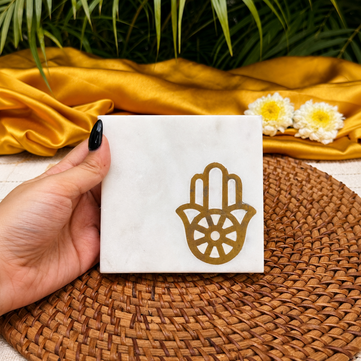 A hand with black nail polish holds a square white marble coaster featuring a gold Hamsa hand design. The coaster is displayed on a brown woven placemat, with a draped gold fabric, white flowers, and green leaves in the background.